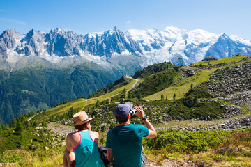 Fototapeta na wymiar French Alps summer travel. Couple of hikers (unidentified people, back view) admire beautiful snow covered Mont Blanc mountains range, resting before keeping to walk. France nature tourism background