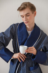 young man in a blue coat stands against a white wall with a cup of coffee