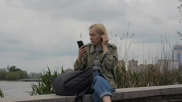 Woman sitting on the banks of the river connect the headphones to the smartphone