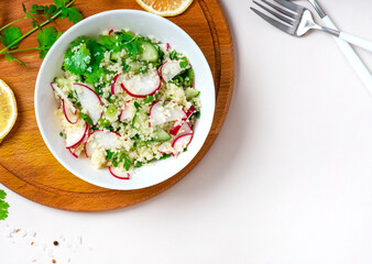 Fresh summer vegetable salad of couscous, radish, cucumbers, parsley and lemon in a bowl on a light background, top view, copy space. Vegan food. Spring or summer food. Free space for text.