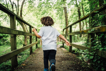4-year-old brown boy running backwards across a bridge in a beautiful green forest with yellow sunlight