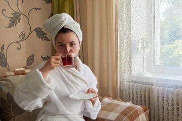 A girl in a white bathrobe is relaxing in a spa on the couch drinking tea.