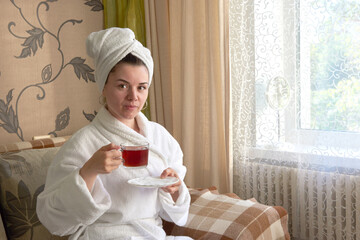 A girl in a white bathrobe is relaxing in a spa on the couch drinking tea.