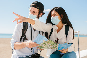 Couple with a mask sitting in front of a beach looking at a map while she points to a direction