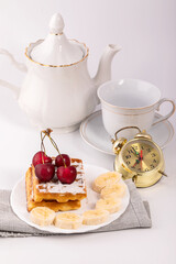 Fototapeta premium Belgian waffles in powdered sugar with bananas and cherries on a white plate. In the background, a teapot and a Cup for morning tea. Delicious breakfast.