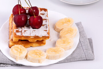 Belgian waffles in powdered sugar with bananas and cherries on a white plate.