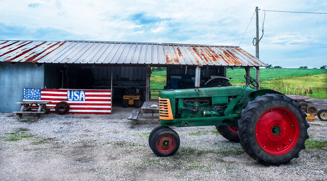 Old antique green farm tractor in front of farm country barn with tin roof and painted American flag