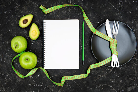 Flat lay of crossed fork and knife tied together with a measuring tape on a black plate, blank paper notebook, wooden pencil, fresh green apples and avocado on black background. Diet journal concept.