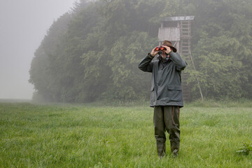 On a rainy day a hunter stands in front of the hunting pulpit in the meadow and looks through his...