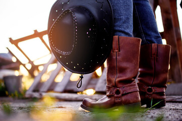 The girl stands on the ranch in cowboy boots and catches the rays of the setting sun.