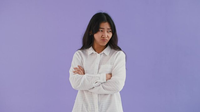 Asian model in white shirt. She has folded her hands and looking offended, shaking head like saying no. Posing against purple background. Close up