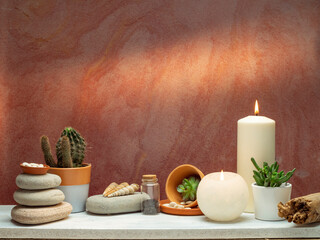 Room decoration with candles, rocks, bottle with black sand and cactus on white shelf against old brick color wall.