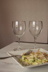 Beautiful glass transparent glasses and white square plates with food stand on a light wooden table

