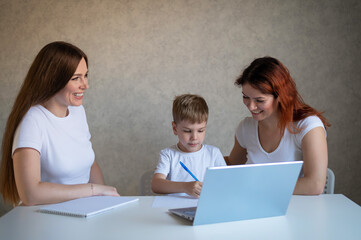 Happy same-sex lesbian family with a child stays at home. Two mothers help the boy do school homework. A couple of women help their son with distance learning. Quarantine.