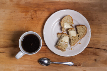 A white Cup with a spoon and coffee, on a wooden Board, a white plate with a slice of homemade bread and butter.