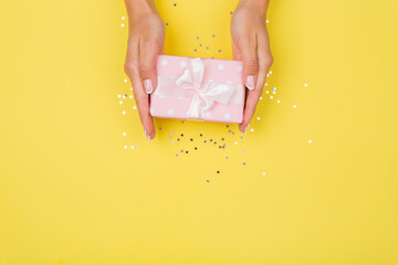 Women hands holding a gift or gift box decorated with confetti on a yellow background top view.