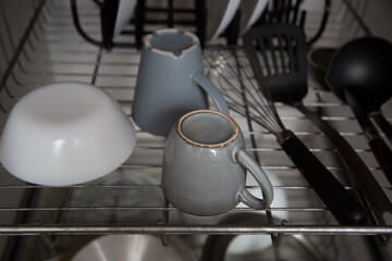gray ceramic cups, a white bowl and black spatula, a whisk and a ladle on a metal grate in a dish dryer