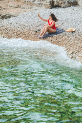 young pretty woman in red swimsuit sitting at rocky beach with phone taking selfie picture