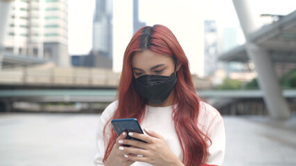 Obraz premium A woman dyed in red hair wearing a mask and standing playing with a mobile phone in the city. 4k Resolution.