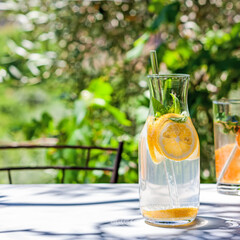 Cold refreshing summer drink or cocktail in carafe with glass straw. Detox water or lemonade with slices of lemon and basil in glass carafe with recyclable straw. Summertime zero wast concept. 