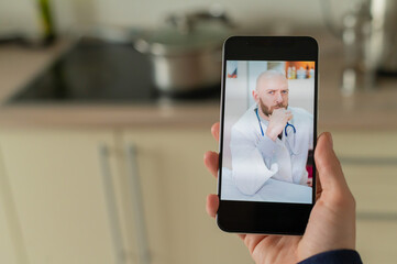 A serious bearded doctor communicates with a patient via a video call on a smartphone. Online medicine concept. Woman in the kitchen takes therapist remote consultation on mobile.