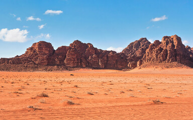 Fototapeta na wymiar Red dusty desert with large rock massif and blue sky in background, small off road vehicle in foreground for scale. Typical landscape of Wadi Rum, Jordan