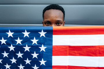 Afro-American guy stands covering his face with the flag of America and looks at the camera