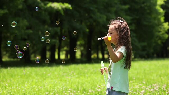 a little girl on a green lawn near the forest blows soap bubbles in the wind