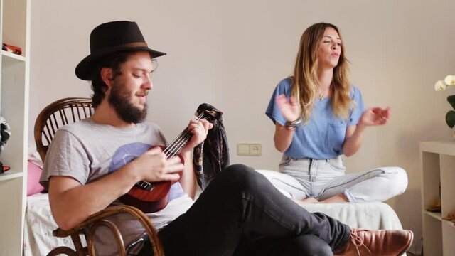 Happy family at home have fan man play ukelele woman sitting on a bad and dancing