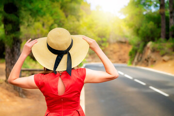 Fototapeta premium Woman in red dress and hat stand by the road in forest on a sunny day.