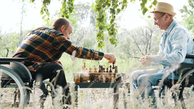 Two happy adult disabled paralyzed friends men sitting in wheelchair play board chess game together in forest. Comfortable leisure, smart activity, fun life of people with physical impairment outdoors