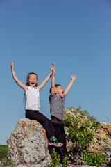 Full body happy boy and girl raising arms and screaming while sitting on boulder against cloudless blue sky on sunny day in countryside