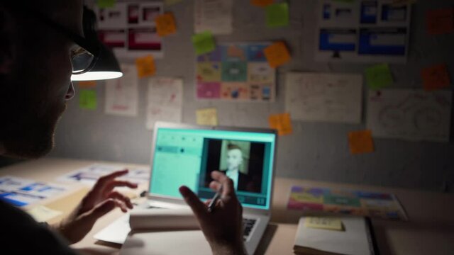 Over the shoulder view of man in eyeglasses having video call with business partner on laptop computer, discussing business sitting at desk lit by lamp at home at night