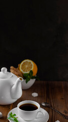 White ceramic teapot, tea set with dark brown hot drink, and natural healthy flavours lemon, lump sugar, mint and ginger on old wooden table near dark wall. Vertical banner, copy space. Rustic style.