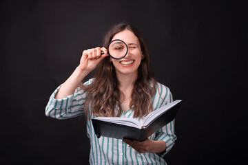 Smiling girl looking through a magnifying glass is holding a journal over black background
