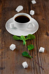white porcelain tea-set with hot drink in on brown rustic wooden table with fresh green mint sprig put together like flower surrounded by pieces of sugar . Creative food concept. Vertical, copy space