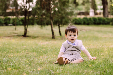 a child sits on a green lawn in a park, a one-year-old boy spends time in the fresh air, a place for text, preschool age, a stylishly dressed child, a piercing look, a child is pondering something