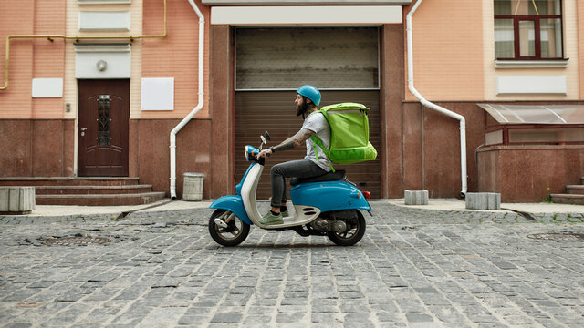 Trendy Food Delivery. Bearded delivery man in helmet with thermo bag or backpack riding a motor scooter along the city, delivering food. Courier, delivery service concept