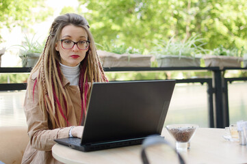 Employed attractive female entrepreneur with long dreadlocks hair is busy writing in her laptop computer while sitting in modern coffee shop outdoors