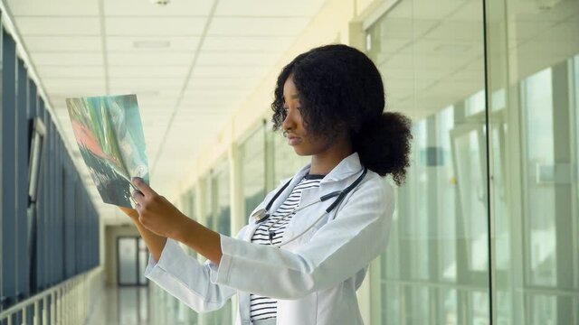 Female african-american doctor examines x-ray of lungs, holding it in hands indoors. Specialist holds transparent image of chest in arms, and carefully researches it