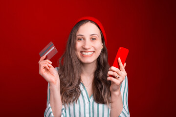 Excited young woman is holding a phone and credit or debit card over red background