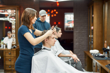 Side view of professional barber girl holding scissors and comb in hands and cutting hair of young guy in the modern barbershop