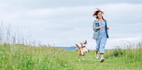 Happy smiling jogging female with fluttering hairs and her beagle dog running and looking at eyes....