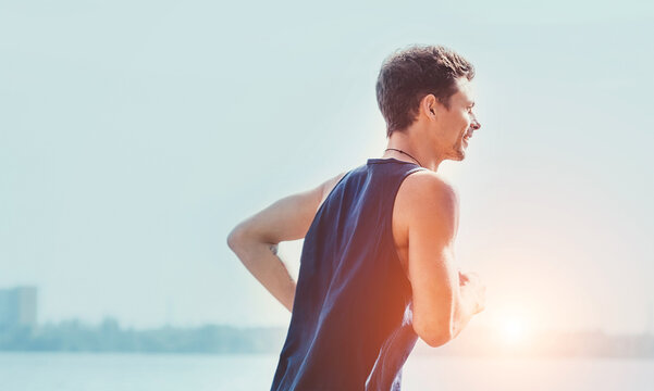 Active athlete muscular young man dressed sleeveless shirt running by the river promenade on the sunny day. He enjoying a jogging swinging hands. Weekend sport activities concept image.