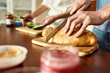 Male hands cutting tasty homemade bread on the wooden board. Young couple preparing healthy meal in the kitchen together. Vegetarianism, healthy food concept.