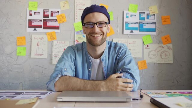 Portrait of handsome young man in eyeglasses and hat working on pc, closing laptop, looking at camera and smiling cheerfully sitting at desk in office or at home surrounded by papers and sticky notes