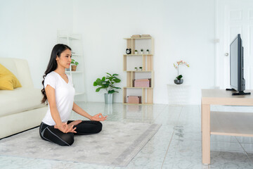 Beautiful young Asian woman in sports clothing doing yoga while relaxing in living room at home to...