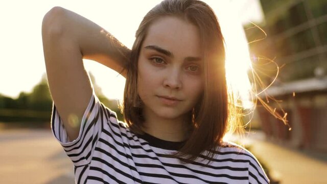 Calm woman touching hair on street during subset and sensually looking at camera