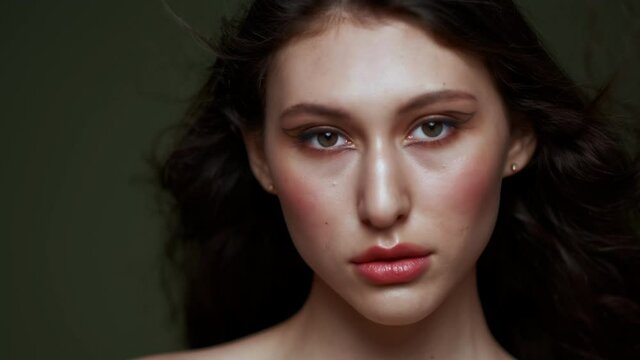 A beautiful young woman with long curly black hair, breast portrait, bare shoulders. Beautiful natural skin. Nude makeup. Red lips and graceful hands. Girl poses in studio against an olive background.