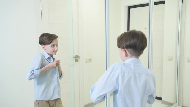 A brunette boy straightens his hair and fastens a blue shirt near the mirror.